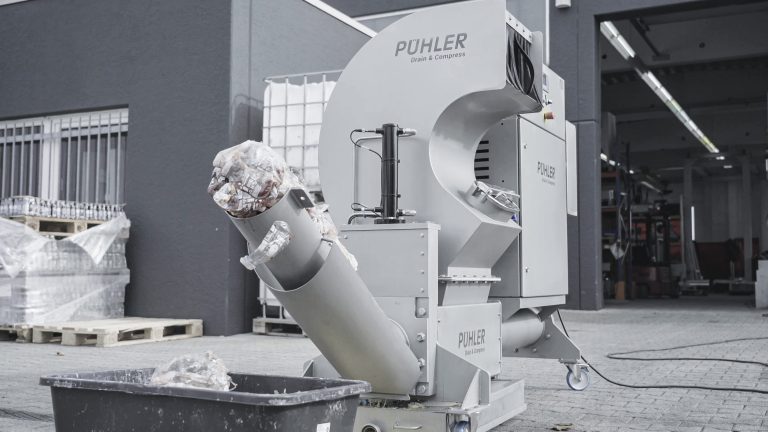 a gray PUEHLER machine in operation, currently ejecting a disc from the discharge. A collection bin is placed below it to collect the discs as they fall from the discharge chute.