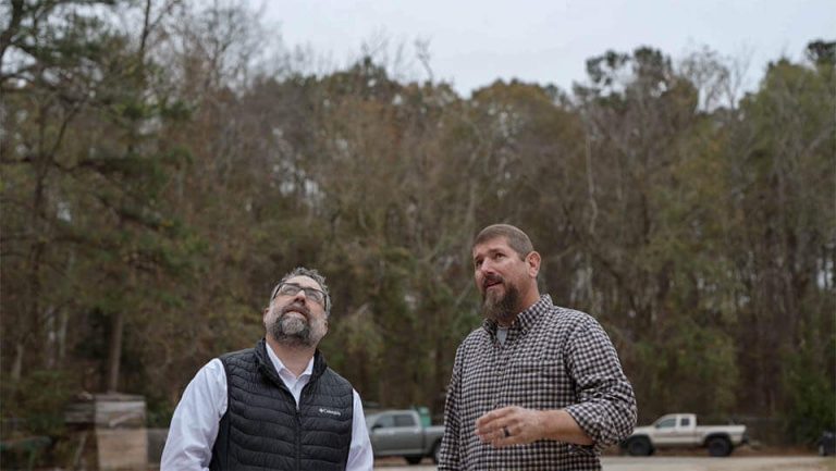 Two people stand next to each other. The woods are behind them. It is a cloudy day.