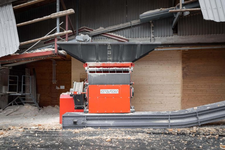A WEIMA S7.15 is standing in front of a building. A conveyor belt with pieces of wood comes from the left. On the right there is a conveyor belt that transports the wood chips.