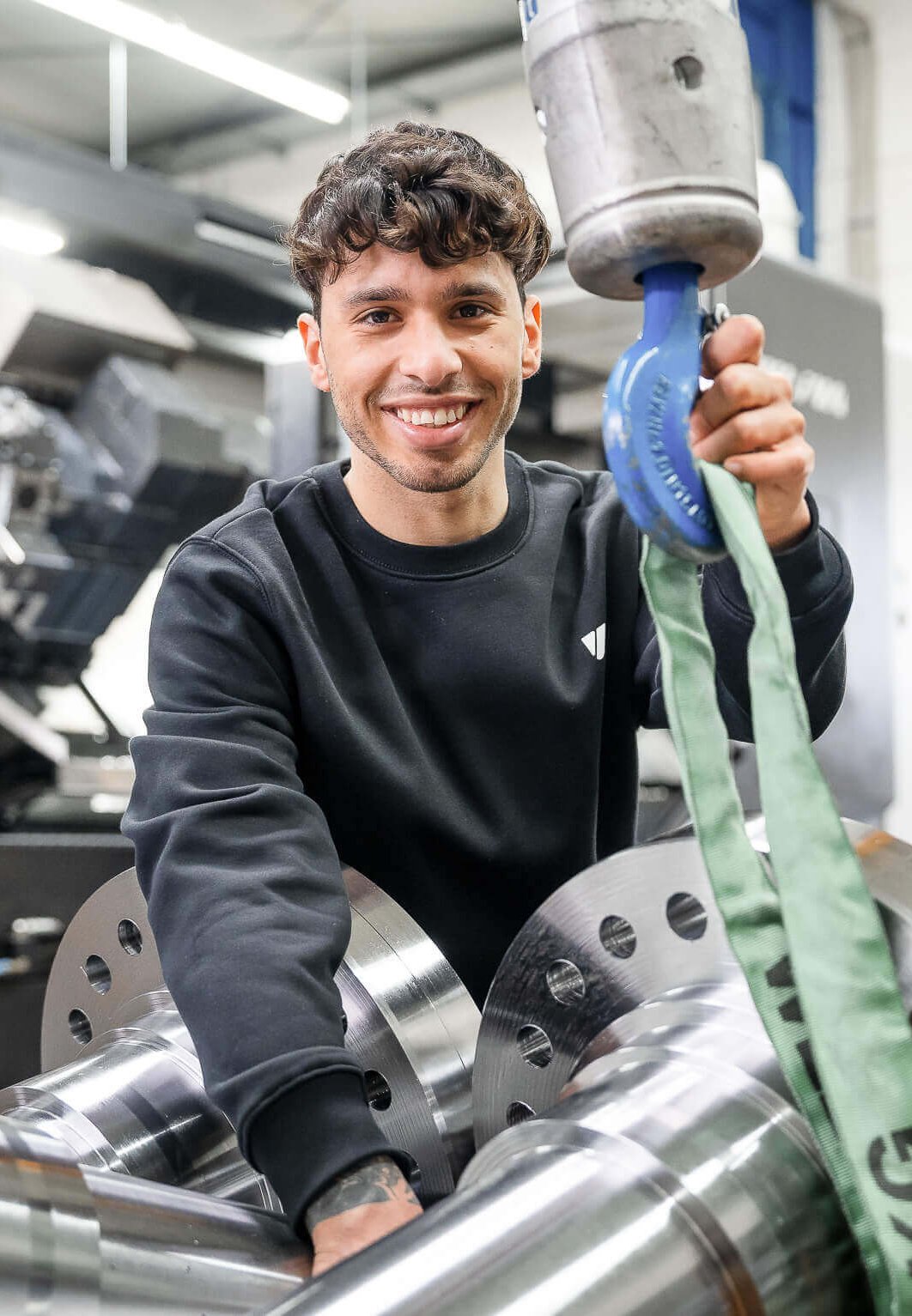 Lächelnder Mitarbeiter vor einem CNC-Fraeszentrum, vor ihm ein Wellenzapfen fuer einen Rotor