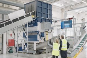 A man and a woman wearing yellow high-visibility vests stand in front of a blue and white shredder that shreds IBCs.
