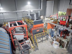 Luftaufnahme eines orangenen Shredders in einer Lagerhalle eingebaut zwischen einem Kran zum befüllen und einem Magnetband