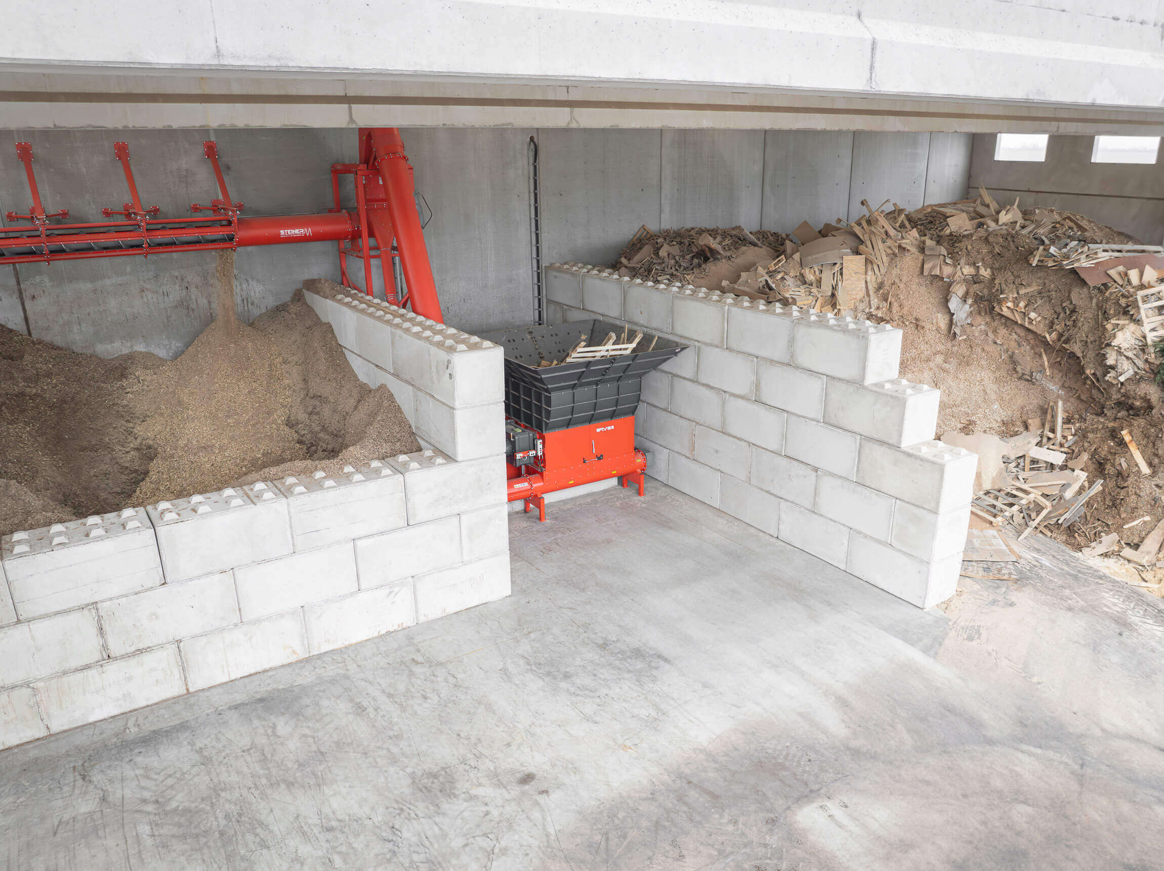 An orange shredder in the middle of large concrete blocks with a screw conveyor between a pile of waste wood and a bunker with wood chips