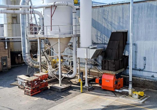 An orange WEIMA shredder is positioned behind a warehouse, surrounded by an air system with a white silo and metal duct work.