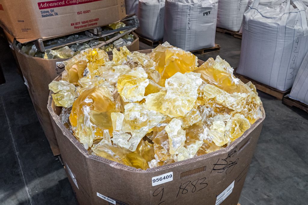 Ein großer Kartonbehälter ist mit gelben, transparenten Kunststoffbrocken gefüllt und steht inmitten weiterer Big Bags in einer Lagerhalle.