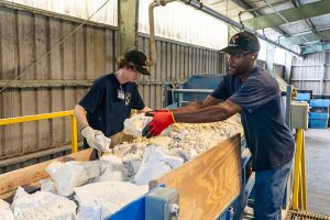 Two workers sort chunks of white material on a conveyor belt in an industrial hall.