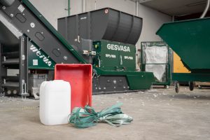 A white canister, a red plastic box and green plastic straps in front of a green shredder for recycling