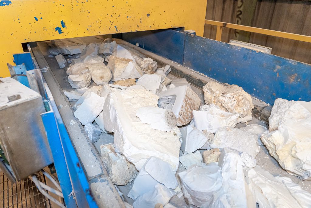 Große weiße und beiges Materialbrocken liegen auf einem Förderband und werden in eine gelbe Industrieanlage zur weiteren Verarbeitung transportiert.