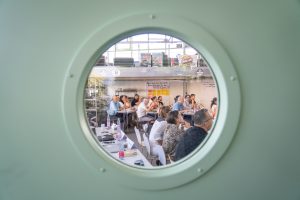 Durch ein Bullauge sieht man die Mitglieder:innen den Vorträgen lauschen