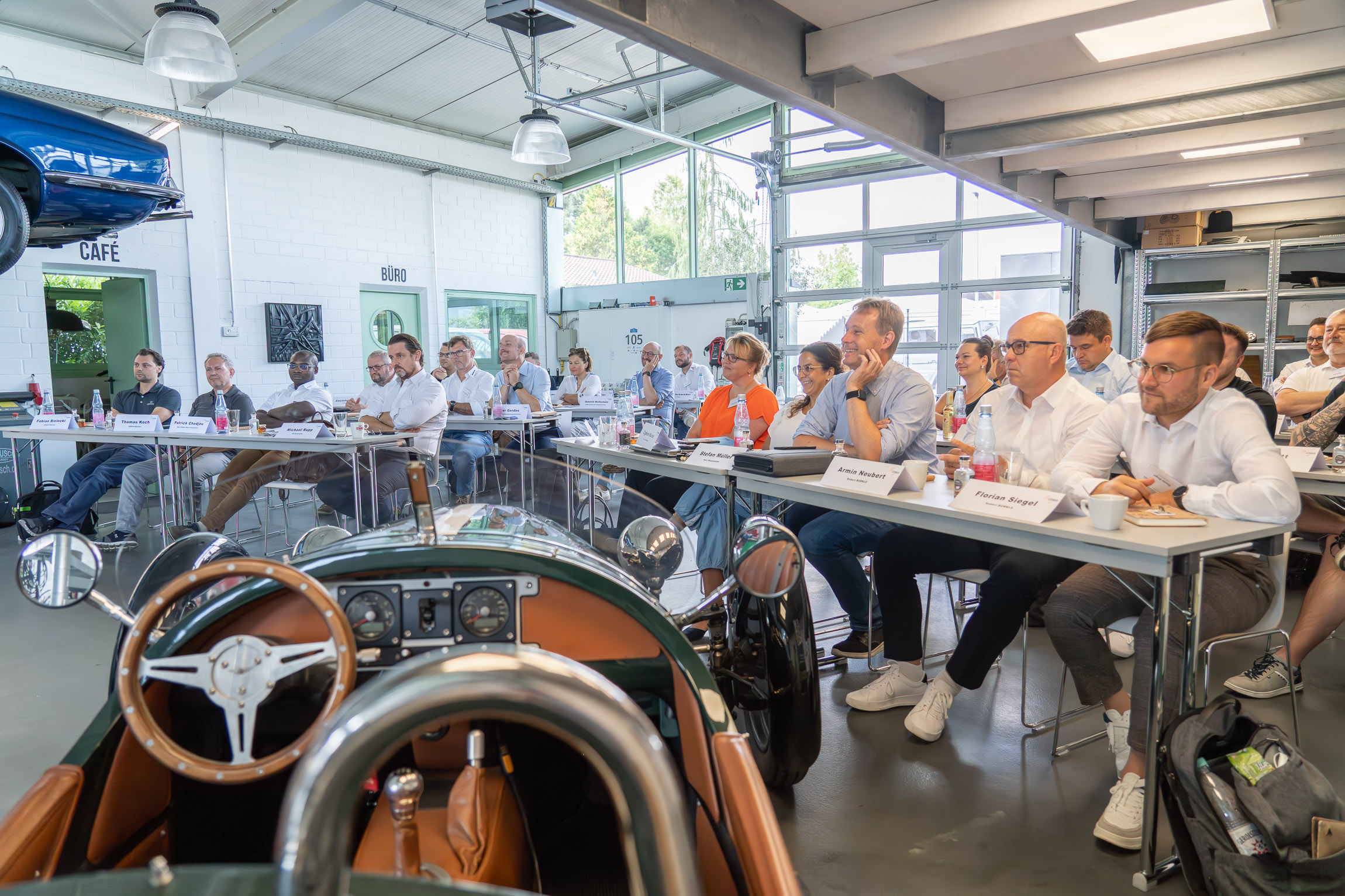 Blick von vorne auf die Mitglieder, die an Konferenztischen sitzen und den Vorträgen zuhören