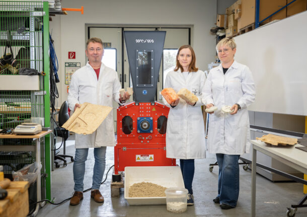 Drei Forscher in weißen Kitteln vor einem orangen Shredder mit verschiedenen Exponaten in der Hand.