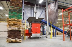 Ein orangefarbener Shredder mit einem Förderband und angeschlossener Absaugung in einer Industriehalle zwischen gestapelten Holzplanken.