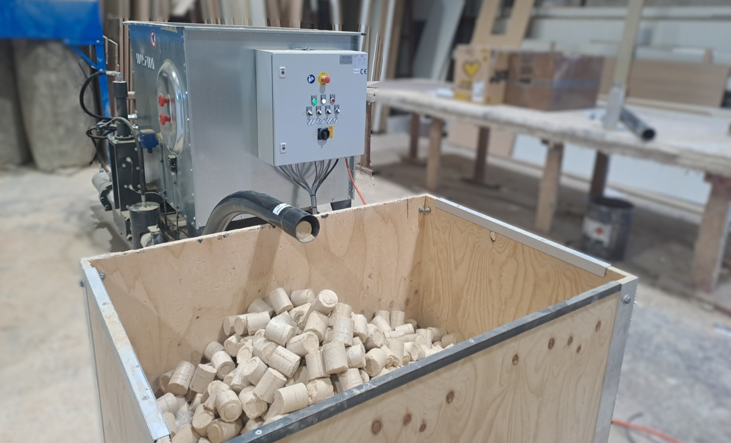 A gray briquetting press behind a box filled with wood dust briquettes.