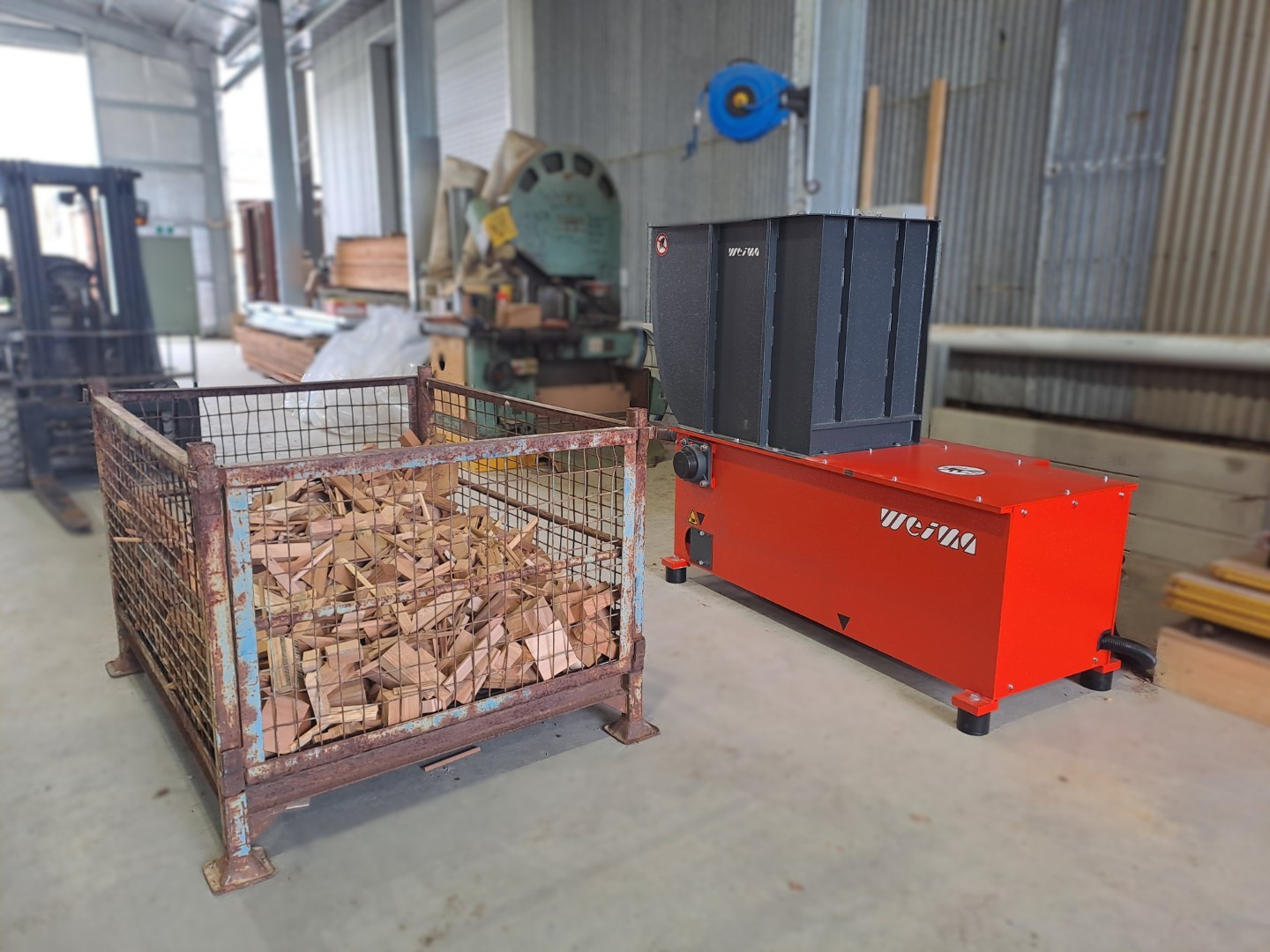 An orange shredder next to a mesh crate filled with wood scraps in a warehouse.