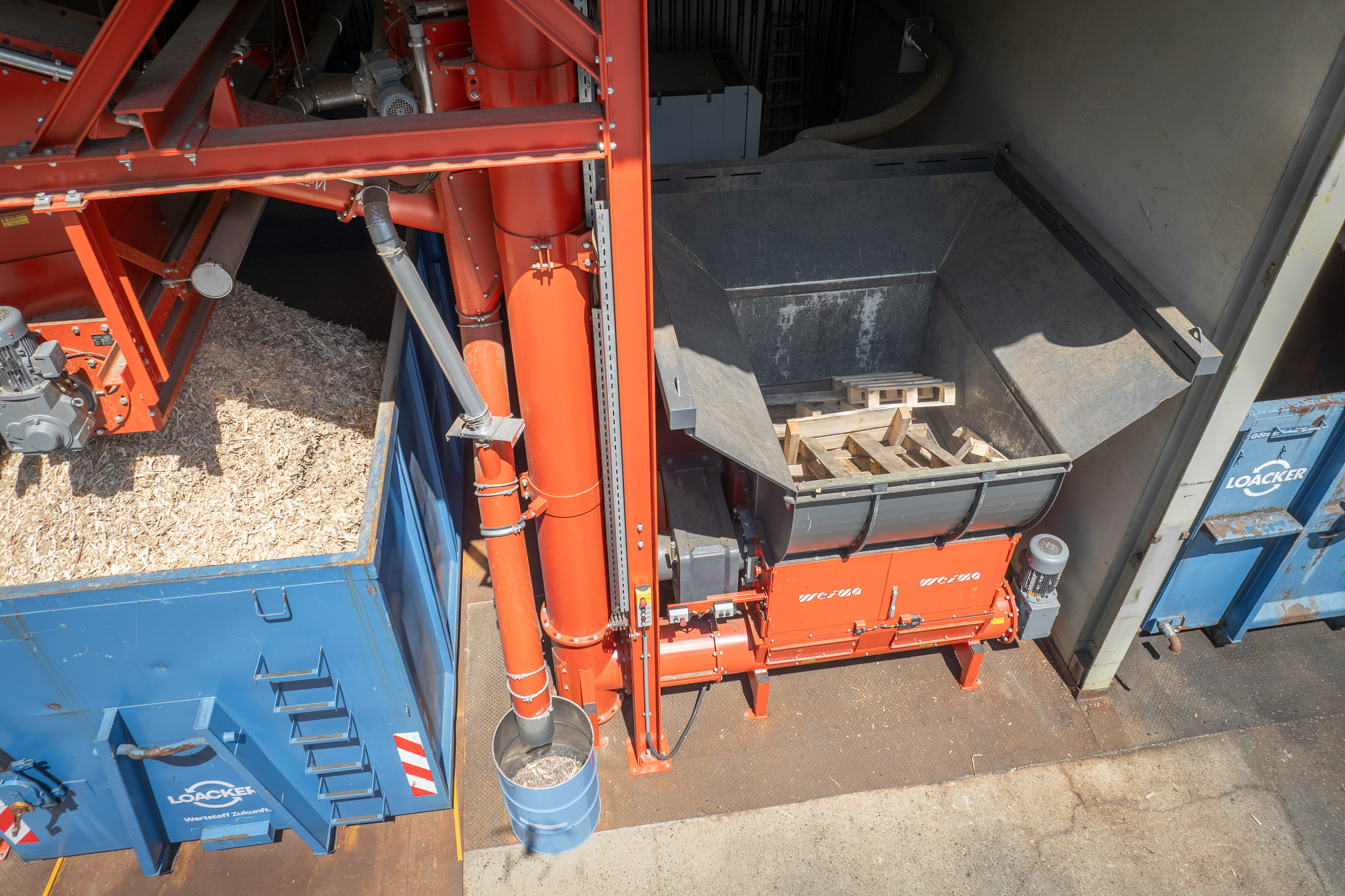 Drone shot of an orange wood shredder in operation, featuring a conveyor system and a container filled with wood chips alongside it.