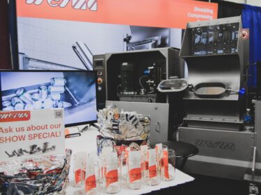 A WEIMA E.200 machine is in a tradeshow booth with a TV and some glasses for giveaways.
