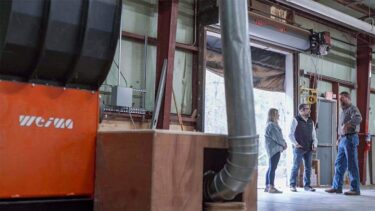 Drei Personen stehen in einer Lagerhalle neben einem blutorangefarbenen WEIMA Shredder und sprechen miteinander.
