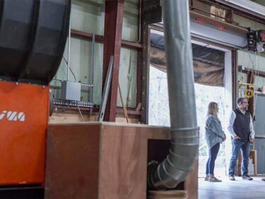 Three people are standing and speaking with each other in a warehouse next to an orange WEIMA shredder.