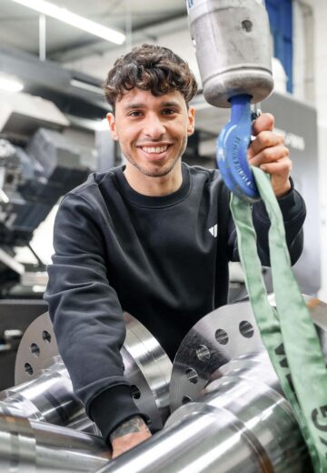 Lächelnder Mitarbeiter vor einem CNC-Fraeszentrum, vor ihm ein Wellenzapfen fuer einen Rotor