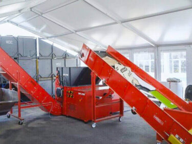A red shredder machine is connected to an infeed and discharge conveyor. Pieces of wood waste are on the infeed conveyor. A black bin waits at the end of the discharge conveyor to collect the shredded material.