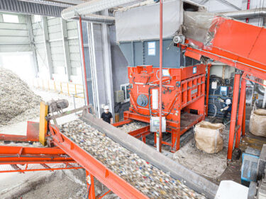 An orange shredder in an industrial plant with a conveyor belt full of shredded waste