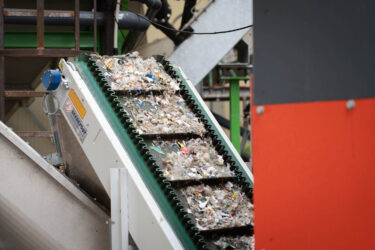 Ein grünes Förderband mit zerkleinerten bunten Post-Consumer Kunststoff mit einem orangenen Shredder im Vordergrund
