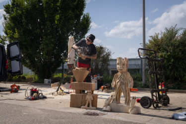 Ein Mann schnitzt mit einer Kettensäge Figuren aus einem Holzblock. Im Vordergrund stehen bereits fertige Figuren