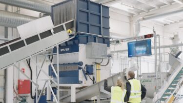 A man and a woman wearing yellow high-visibility vests stand in front of a blue and white shredder that shreds IBCs.