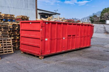 A red dumpster behind a warehouse is filled with wood scraps. Next to it is a stack of wooden pallets.