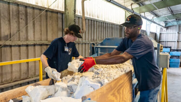 Two workers sort chunks of white material on a conveyor belt in an industrial hall.