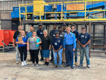 A group of employees stand together for a group photo in an industrial hall in front of a recycling plant.
