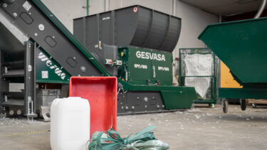 A white canister, a red plastic box and green plastic straps in front of a green shredder for recycling