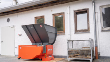 Ein orangefarbener Shredder vor einem weißen Gebäude mit einer Kiste mit alten Fassdauben