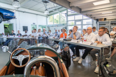 Blick von vorne auf die Mitglieder, die an Konferenztischen sitzen und den Vorträgen zuhören