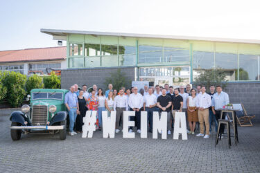Eine Gruppe Personen steht vor einem Gebäude. im Vordergrund sind weiße Buchstabe mit dem Schriftzug #WEIMA, daneben steht ein grüner Oldtimer.