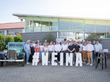 Eine Gruppe Personen steht vor einem Gebäude. im Vordergrund sind weiße Buchstabe mit dem Schriftzug #WEIMA, daneben steht ein grüner Oldtimer.
