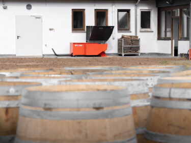 Im Hintergrund ein orangener Shredder von WEIMA, im Vordergrund Barrique-Fässer