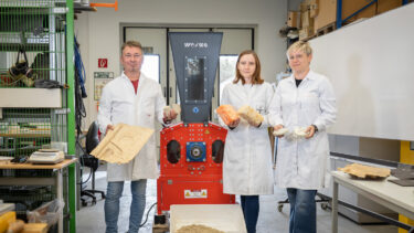Drei Forscher in weißen Kitteln vor einem orangen Shredder mit verschiedenen Exponaten in der Hand.
