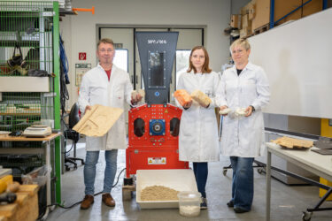 Drei Forscher in weißen Kitteln vor einem orangen Shredder mit verschiedenen Exponaten in der Hand.