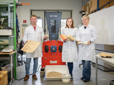 Drei Forscher in weißen Kitteln vor einem orangen Shredder mit verschiedenen Exponaten in der Hand.