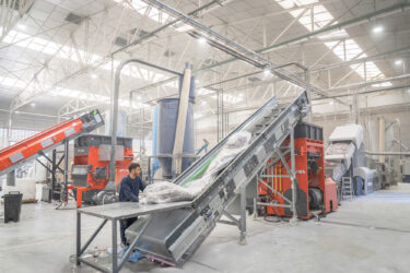 In a hall, you can see a worker manually filling the conveyor belt in front of the shredder with plastic film. In the background, you can see the second shredder