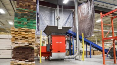 Ein orangefarbener Shredder mit einem Förderband und angeschlossener Absaugung in einer Industriehalle zwischen gestapelten Holzplanken.