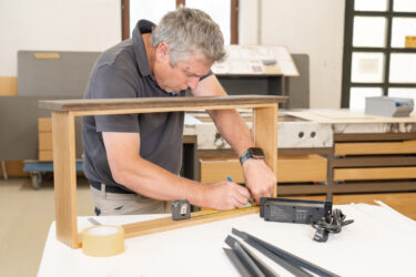 Mitarbeiter in einer Werkstatt, der an einem Holzelement Maß nimmt und Markierungen anzeichnet; auf dem Arbeitstisch liegen Werkzeug und weitere Bauteile