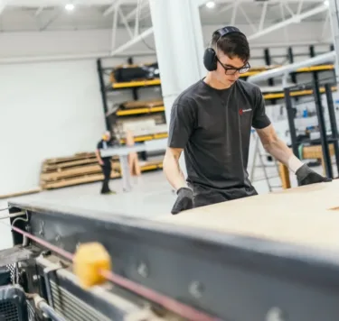 An employee is standing in a hall, placing pieces of wood onto the conveyor belt