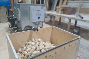 A gray briquetting press behind a box filled with wood dust briquettes.